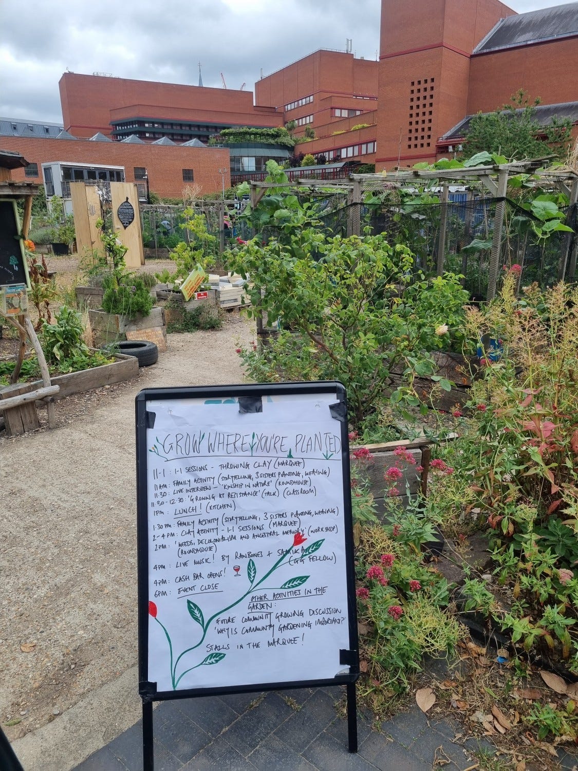 The British library n the background, a red brick garden. in the image, there are lots of small communty garden plots, and a billboard saying grow where you are, with an agenda of talks.