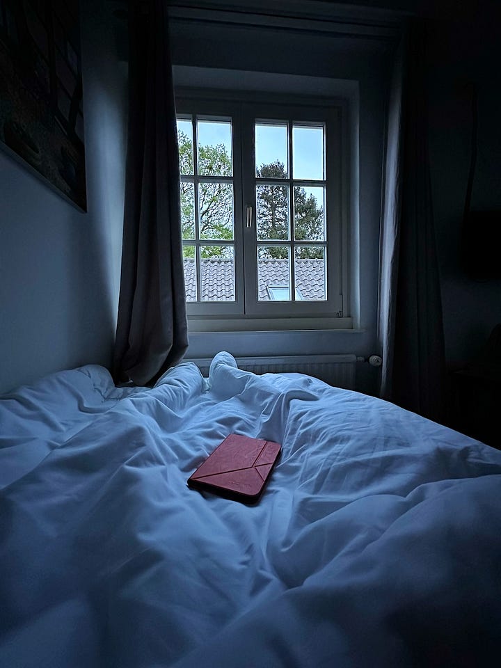 Een e-reader en een stuk vlaai op een terrastafel en een oranje e-readerhoes op een wit dekbed in een halfdonkere kamer.