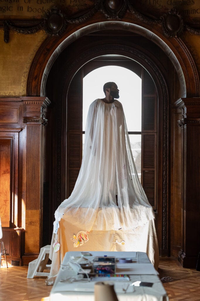 Jerron Herman, a Black man with close-cropped hair and a beard, stands on a platform in a large, wood-paneled room, gazing over his left shoulder. He wears a sheer white cape-like garment that drapes from his shoulders down past his feet. Jerron Herman, a Black man with close-cropped hair and a beard, stands on a platform in a large, wood-paneled room, gazing over his left shoulder. He wears a sheer white cape-like garment that drapes from his shoulders down past his feet.