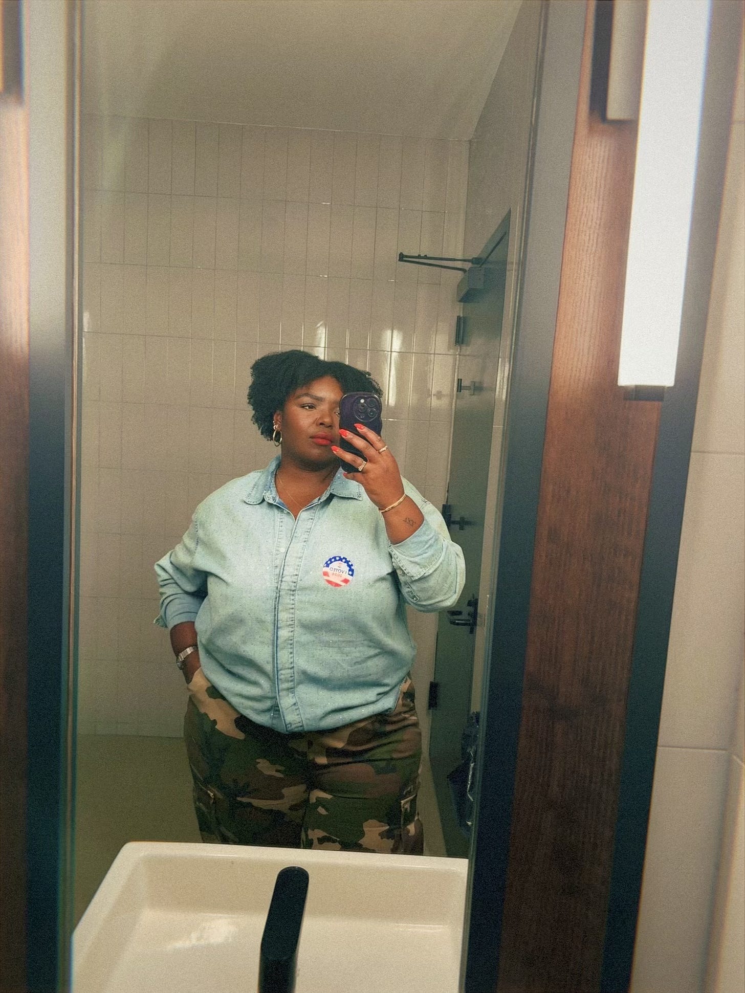 Kamaria mirror selfie, wearing denim top with an "I voted" sticker and came pants.