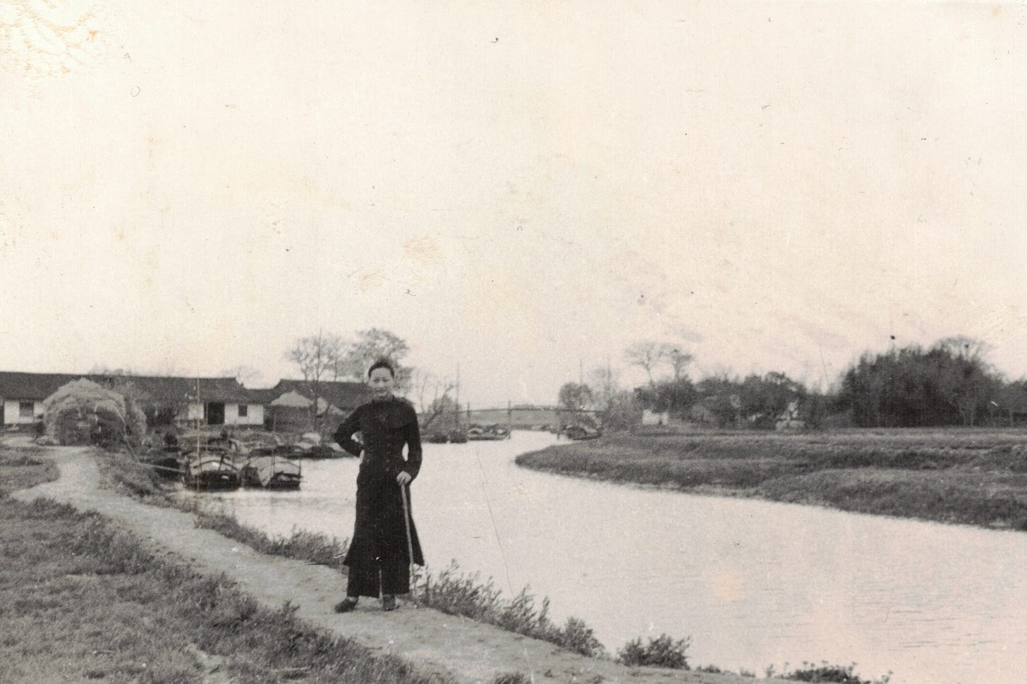 Anna May Wong in all black stands on a dirt path near a canal with farmhouses in the background, rural China Anna May Wong in all black stands on a dirt path near a canal with farmhouses in the background, rural China
