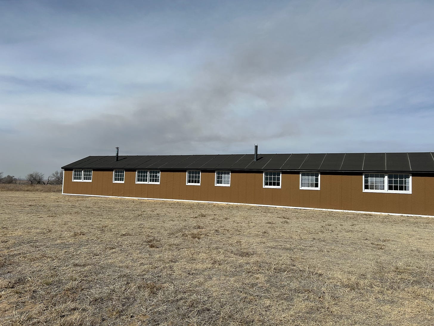 A recreation of one of the Amache residential buildings. The building is long and narrow. Its exterior is a dark brown with windows and a black roof. The squat building is reminiscent of a barn or large-scale turkey shelter. The foreground is brown, dry, dead grasses. The background is a clouded and slightly hazy morning sky.