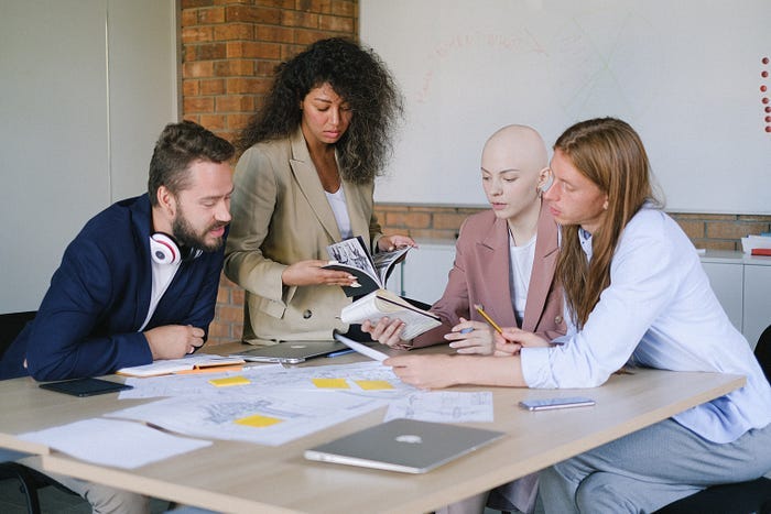 A woman guiding a conversation while 3 other people are looking on, at notebooks, diagrams, and more. A woman guiding a conversation while 3 other people are looking on, at notebooks, diagrams, and more.