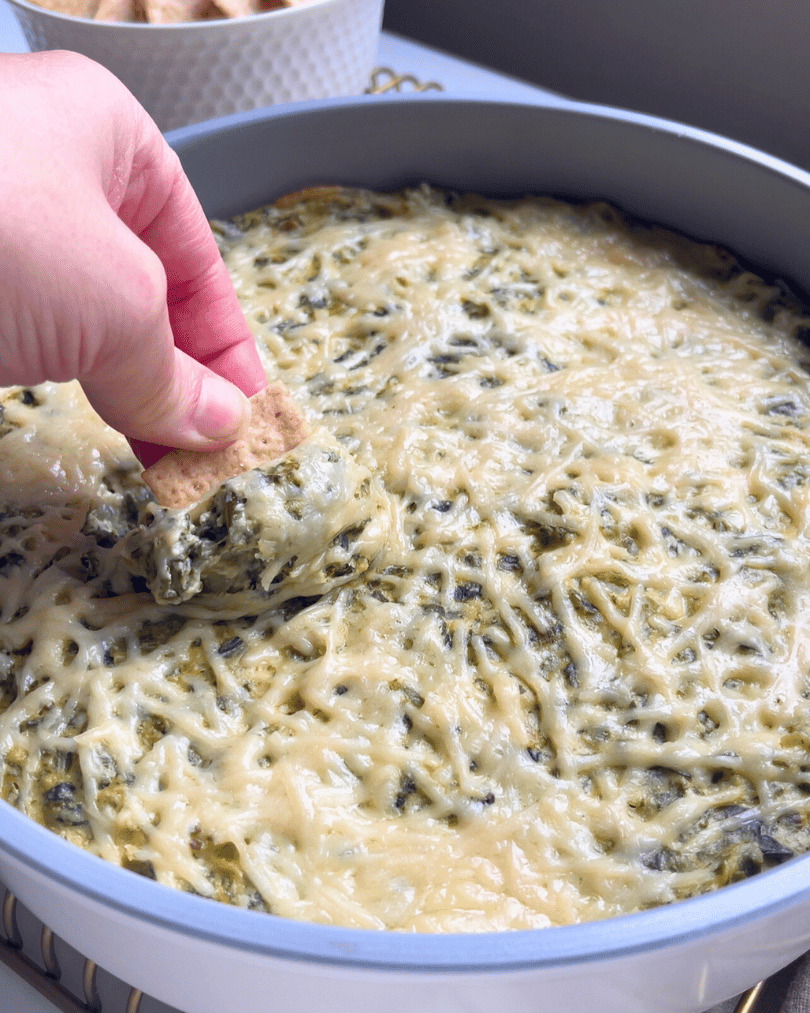vegan spinach artichoke dip on a wire cooling rack with a hand dipping a cracker into it