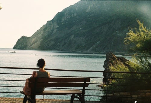 This may contain: a woman sitting on top of a wooden bench next to a body of water with mountains in the background