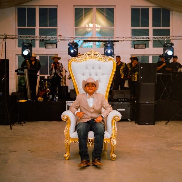 Hermelindo Jaimes, con sombrero vaquero, americana beige y pajarita, sentado en una silla con forma de trono, con una banda en directo detrás de él.