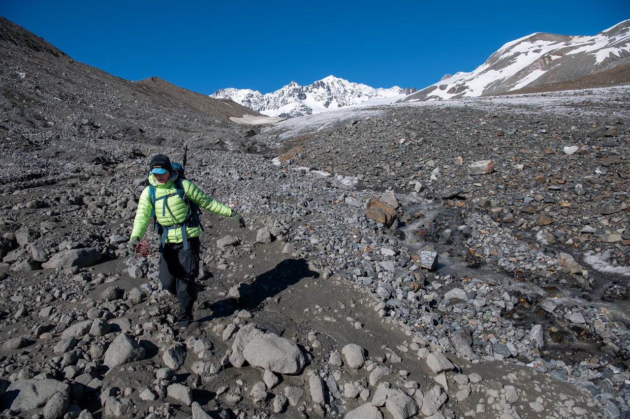 A hiker in a bright green jacket walks down a broad slope of grey rock debris and grimy remnant snow, trekking gear in hand, casting a long shadow. Behind, a dirty snowfield sweeps up to a row of snow-plastered peaks and dark spires against a deep blue sky.
