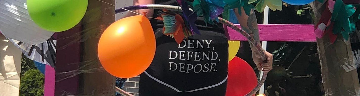 A person holding a hula hoop and wearing booty shorts printed with “DENY, DEFEND, DEPOSE.” The float they’re standing on is decorated with balloons. A person holding a hula hoop and wearing booty shorts printed with “DENY, DEFEND, DEPOSE.” The float they’re standing on is decorated with balloons.