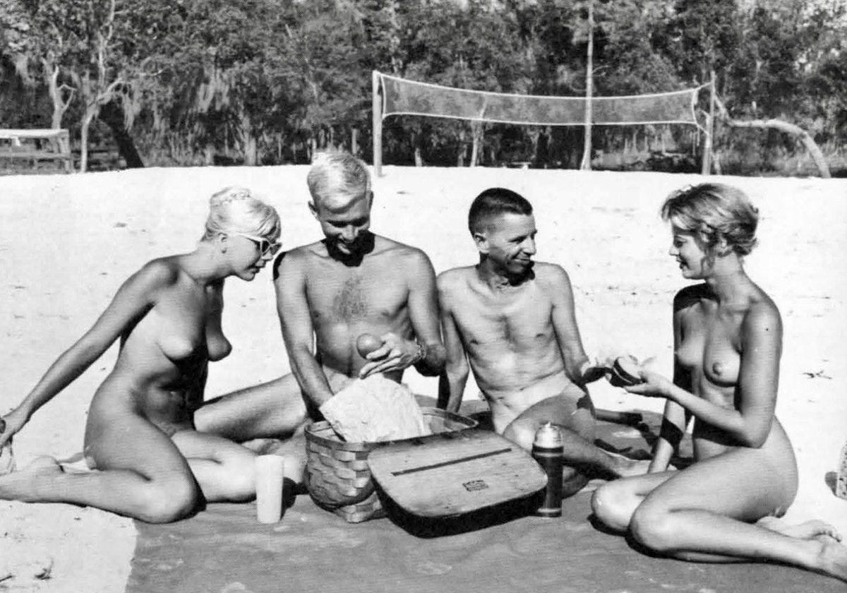 A grainy black and white photo of four nude adults having a picnic on a white florida beach, a volleyball net in the background. 