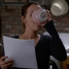 This may contain: a woman drinking from a coffee cup while holding a piece of paper in her hand