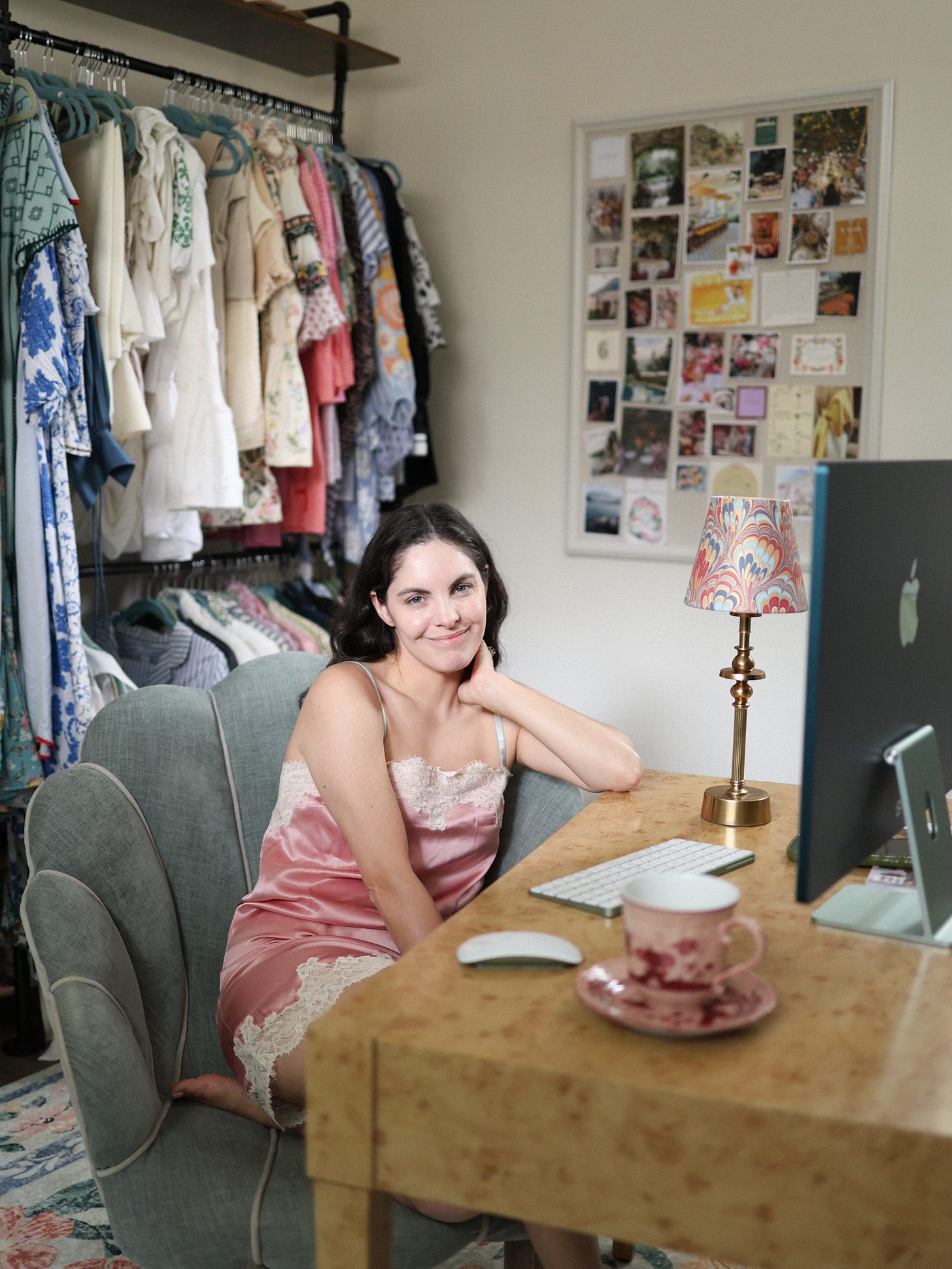 Monica with serene expression seated at wooden desk in her closet office, morning light casting warm glow, with shell-shaped chair, coffee setup, and inspiration board visible behind her.