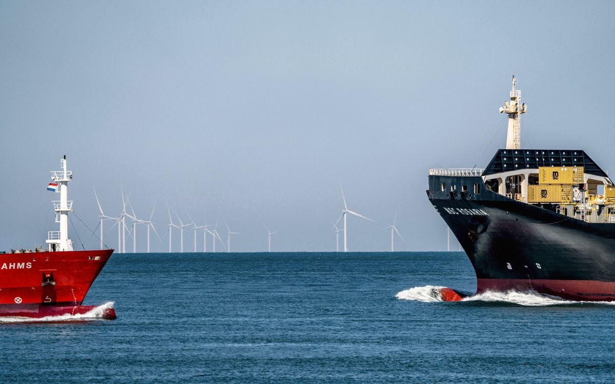 A freighter and container ship crossing paths at sea, with offshore wind turbines in the distant background. A freighter and container ship crossing paths at sea, with offshore wind turbines in the distant background.