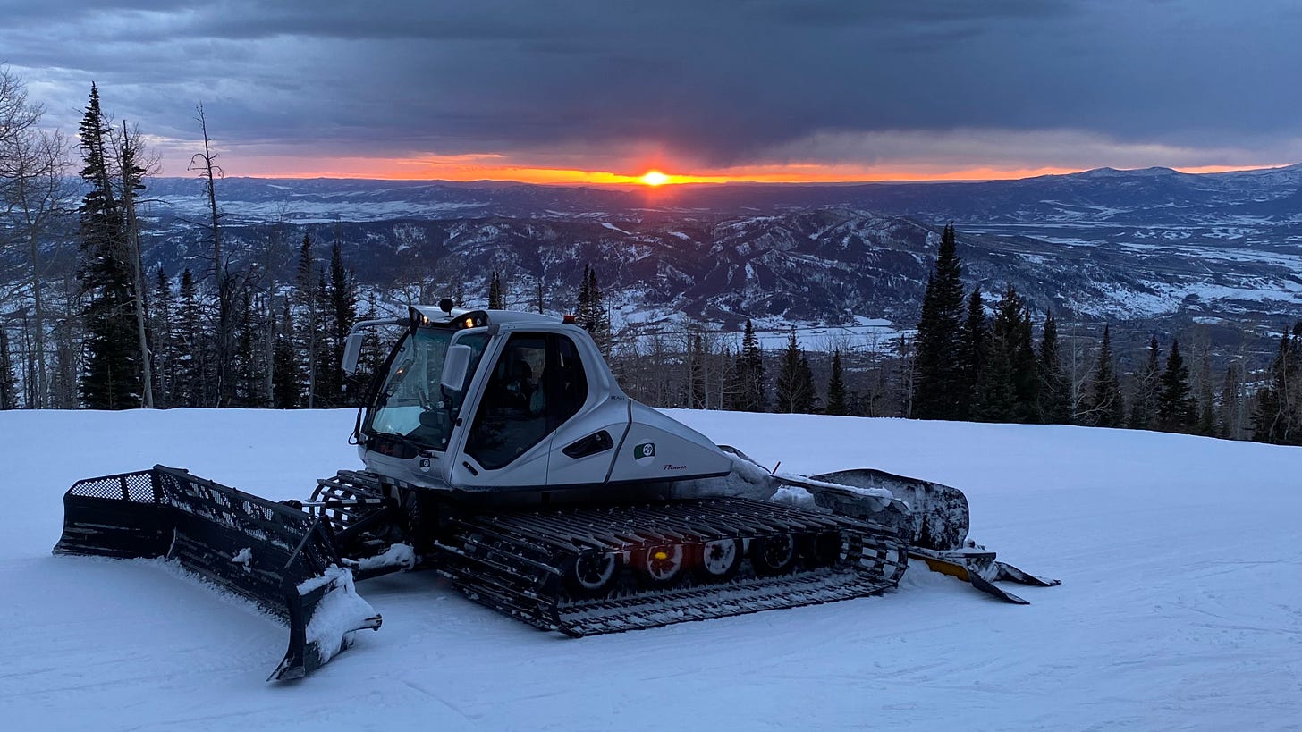 Photo of a snow groomer parked on the top of a mountain with a valley view and sunset in the background.