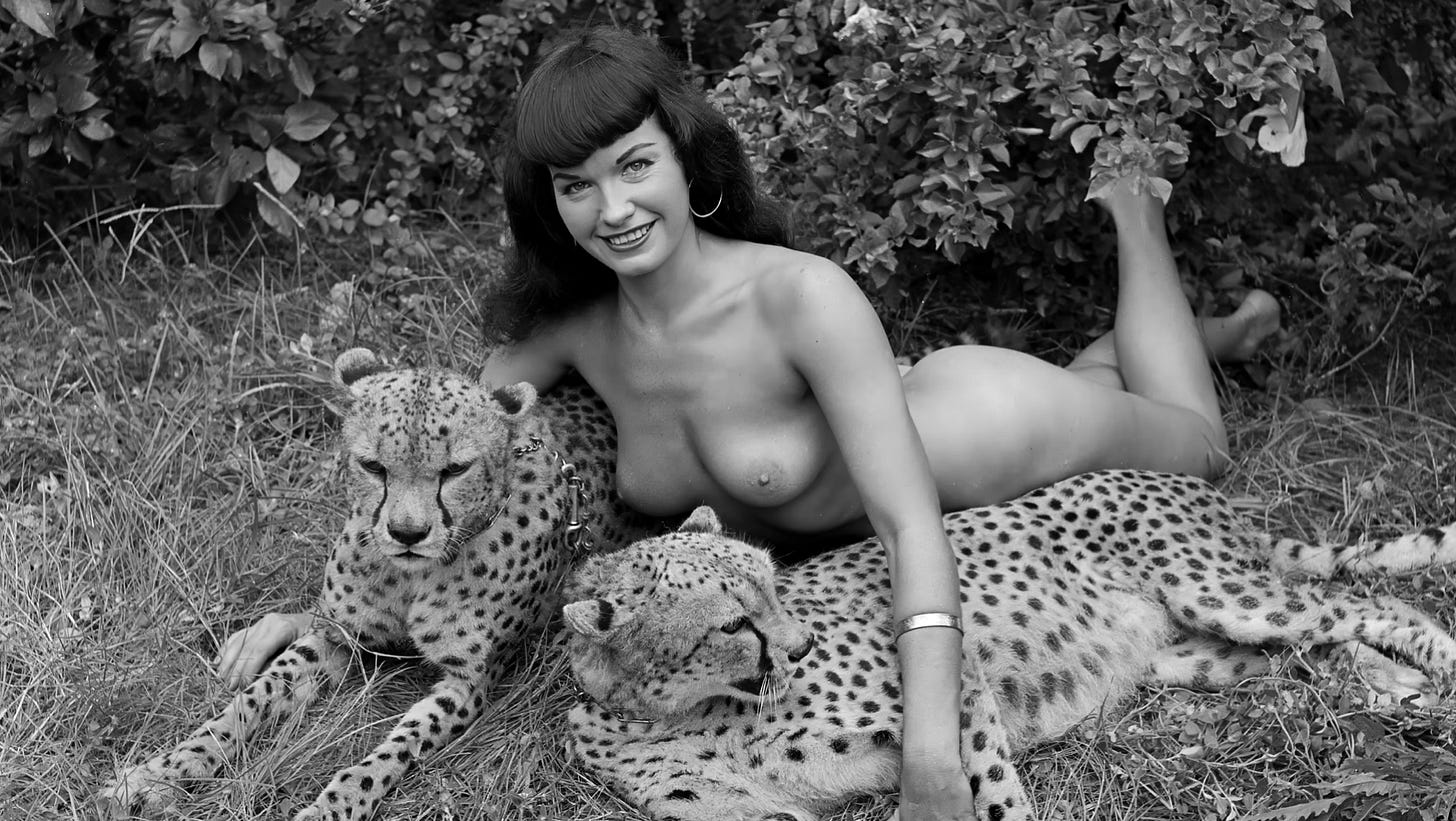 Bettie Page lying nude on the grass, smiling at the camera while resting alongside two calm, spotted cheetahs, with leafy bushes behind them.