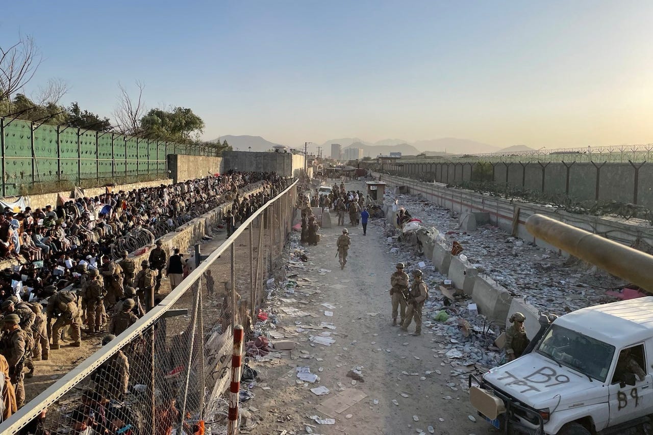 Over a dozen service members in camouflage and hundreds of evacuees are on one side of a chain link fence while a handful of other camouflaged service members patrol on the opposite side of the fence. Over a dozen service members in camouflage and hundreds of evacuees are on one side of a chain link fence while a handful of other camouflaged service members patrol on the opposite side of the fence.