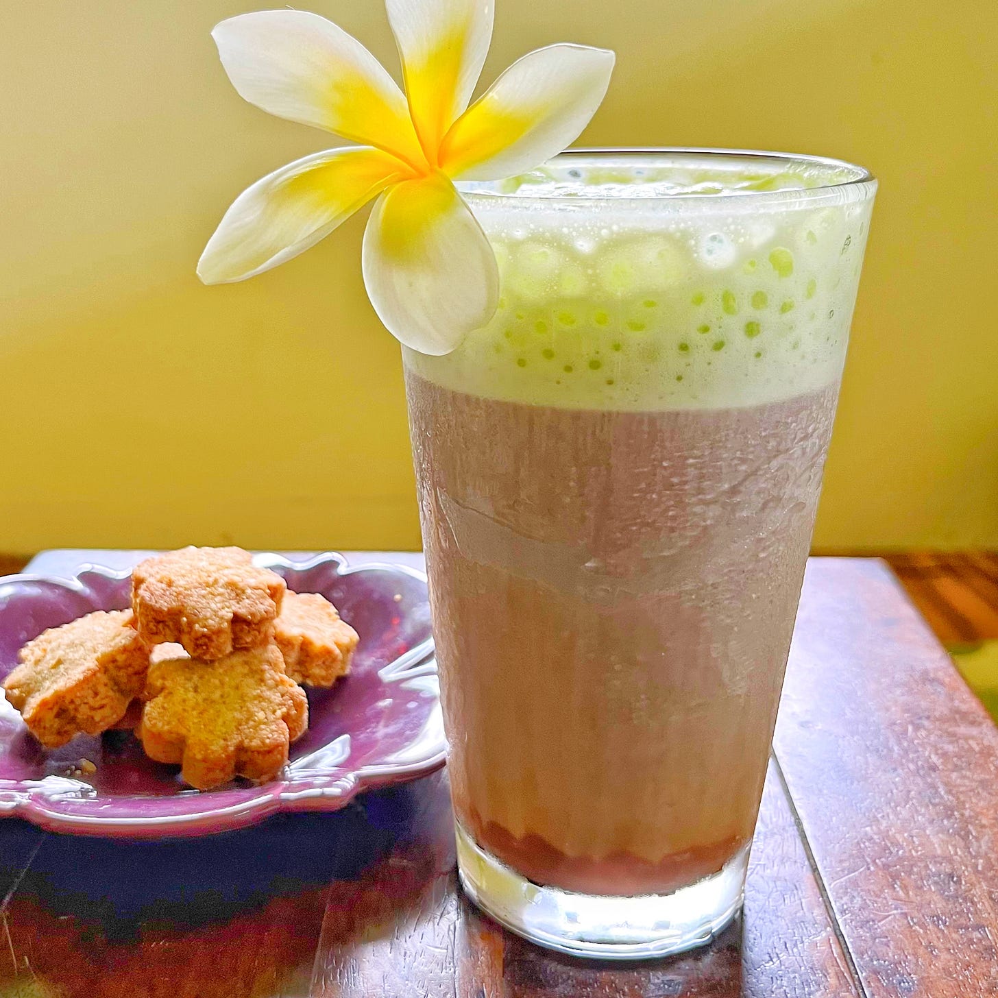 Iced chocolate with green foam in glass with plumeria garnish. Plate of cookies in background