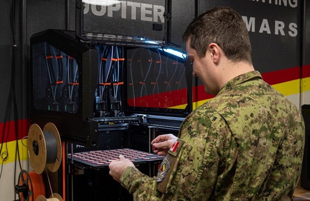 A person wearing a military uniform stands in front of a large industrial 3D printer. The individual is holding a rectangular tray filled with evenly spaced red circular objects. Multiple spools of filament are mounted on the side of the machine. The background wall features bold horizontal stripes in red, yellow, and black, along with large white text partially visible.