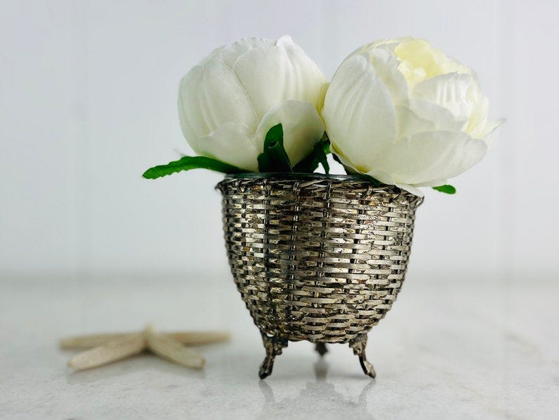 May include: Two white flowers in a silver, footed, woven basket.