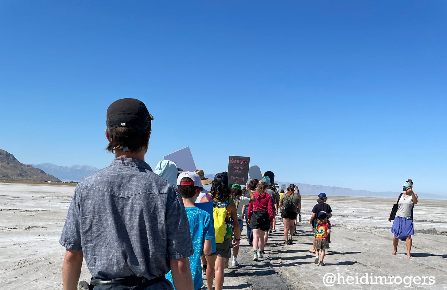 Photo of people from behind, marching in a line on sand toward the Great Salt Lake, with blue sky and low mountains in the distance.