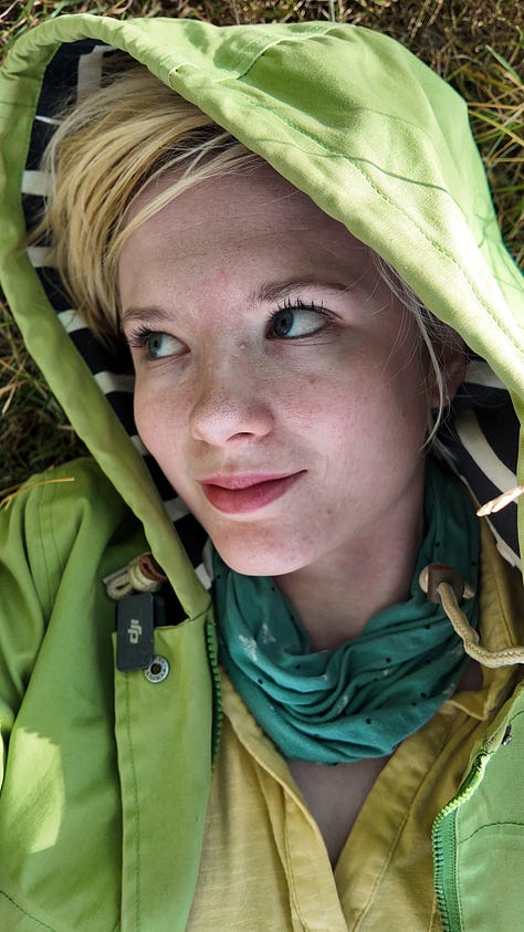 A giant beech tree, Yggdrasil; Lucy, a white woman with a green hooded jacket and blond hair looking to the side, a hand holding a  golden poplar leaf
