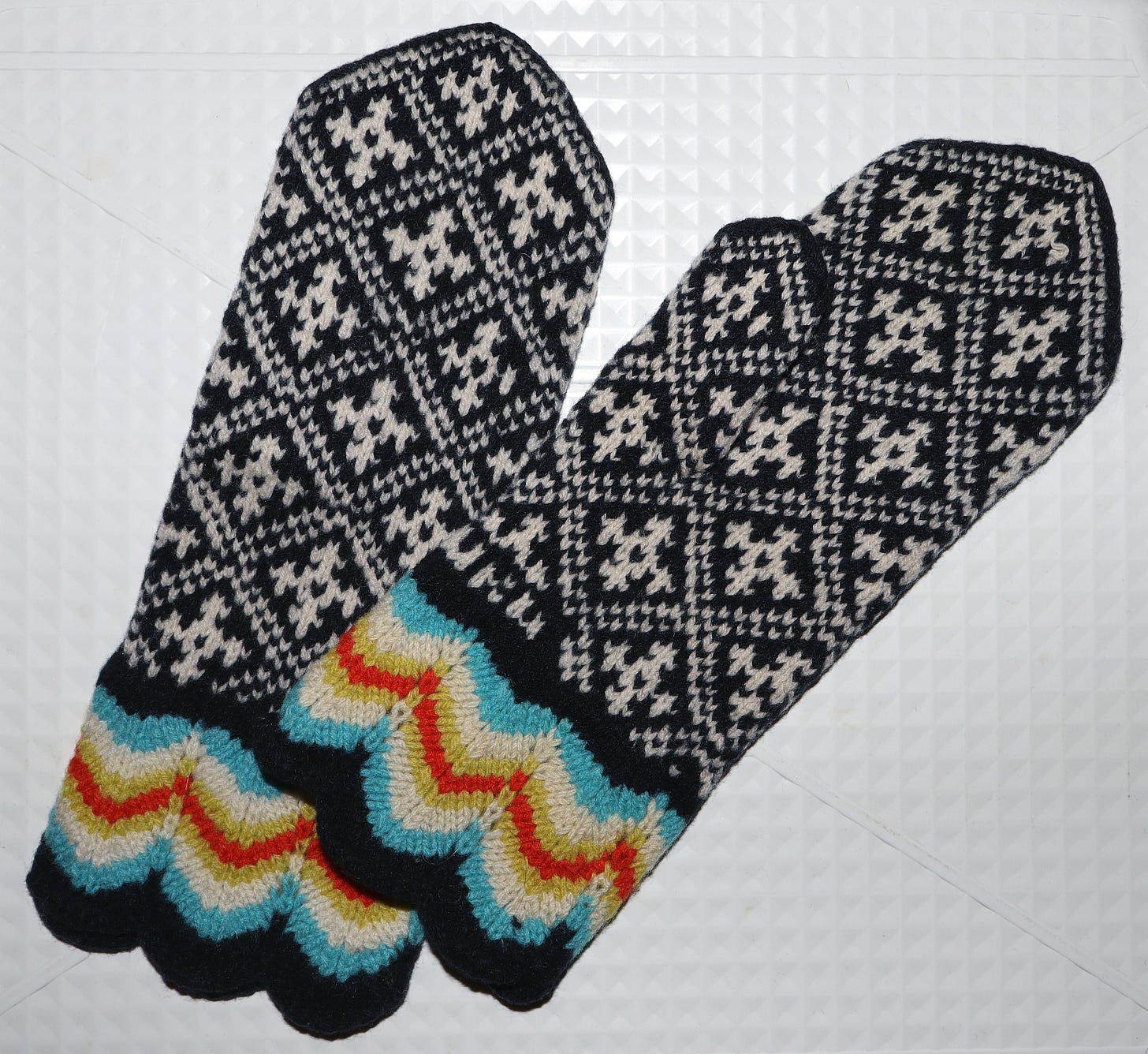 A pair of traditional Estonian mittens laid flat on a white background. The body is black and cream stranded colourwork with repeating geometric cross and diamond patterns. The cuffs feature a colourful zigzag border in red, teal, yellow, and orange on a black ground.