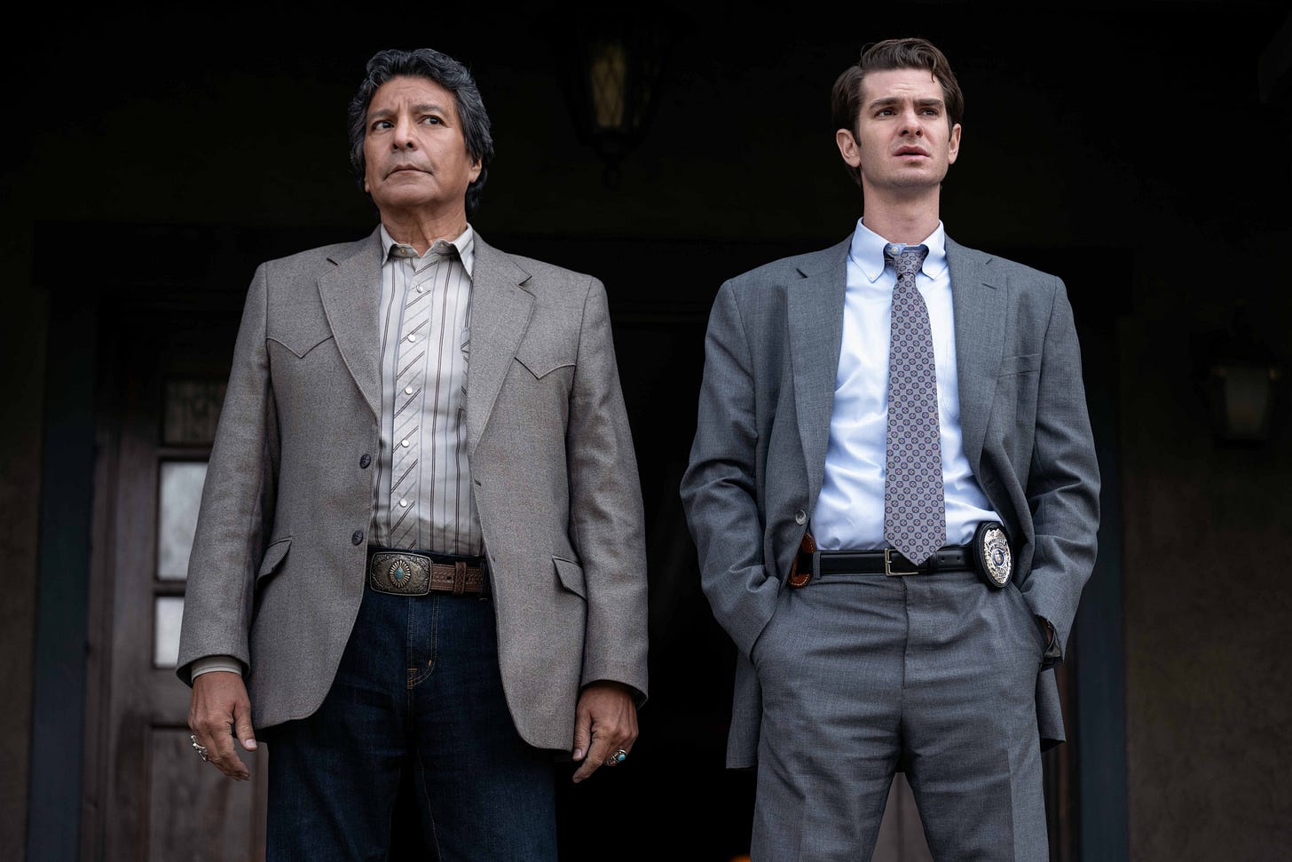 Two men stand side by side on what is apparently the front porch of a wooden house, the camera looking slightly up at them. On the left is an older Native American man dressed neatly in a Western-style sport coat, fancy belt and jeans. On the right stands a young white man dressed in a gray suit and tie. A badge is visible on this man's belt. Two men stand side by side on what is apparently the front porch of a wooden house, the camera looking slightly up at them. On the left is an older Native American man dressed neatly in a Western-style sport coat, fancy belt and jeans. On the right stands a young white man dressed in a gray suit and tie. A badge is visible on this man's belt.