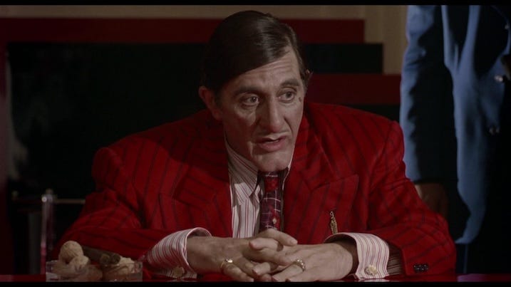 Close-up of Big Boy Caprice (Al Pacino) in a red pinstripe suit sitting at a table, lit with sharp shadows and saturated color—a classic example of the film’s bold production design.
