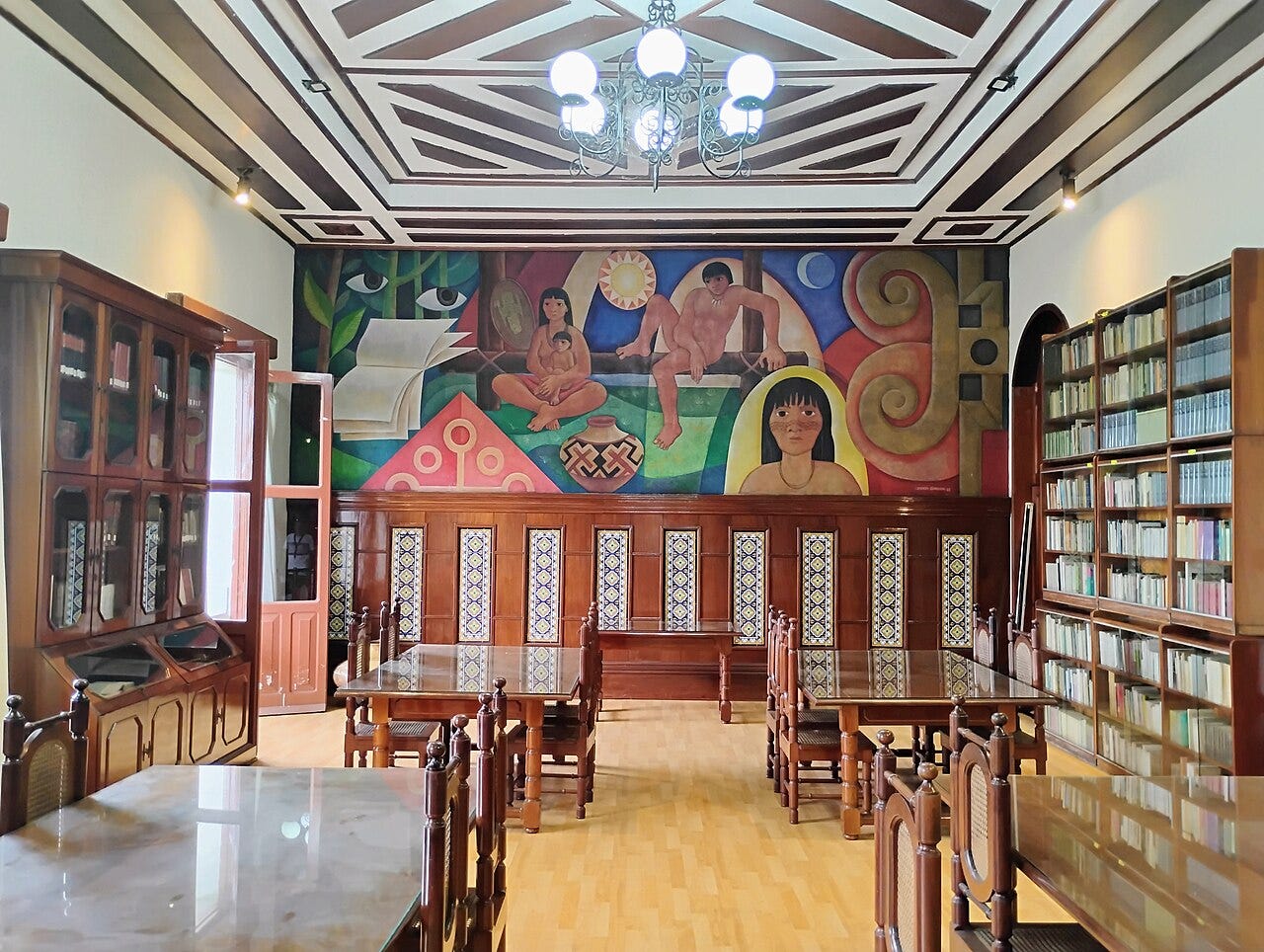 #descrevemospravocê  A imagem mostra uma sala de biblioteca com móveis antigos de madeira e decoração artística vibrante. No fundo da sala, um grande mural colorido ocupa toda a parede. A pintura retrata figuras humanas em tons terrosos, com destaque para uma mulher sentada com uma criança no colo, um homem deitado em uma estrutura de madeira e o rosto de uma jovem com cabelos pretos e franja. Ao redor das figuras, há elementos geométricos, formas abstratas e símbolos que remetem à cultura indígena e latino-americana. No canto esquerdo do mural, uma folha de papel branco flutua no ar diante de olhos verdes em meio à vegetação estilizada.  O teto da sala é de madeira clara com detalhes escuros em padrões diagonais. Um lustre de ferro com lâmpadas brancas pendura-se ao centro. Nas laterais da sala, estantes envidraçadas e prateleiras de madeira contêm livros e documentos. Mesas retangulares com tampo de vidro e cadeiras de encosto alto completam o ambiente, organizado e silencioso. O piso é de madeira clara. A iluminação natural entra por janelas amplas à esquerda da imagem, criando um ambiente acolhedor, que mistura arte, cultura e preservação do conhecimento.
