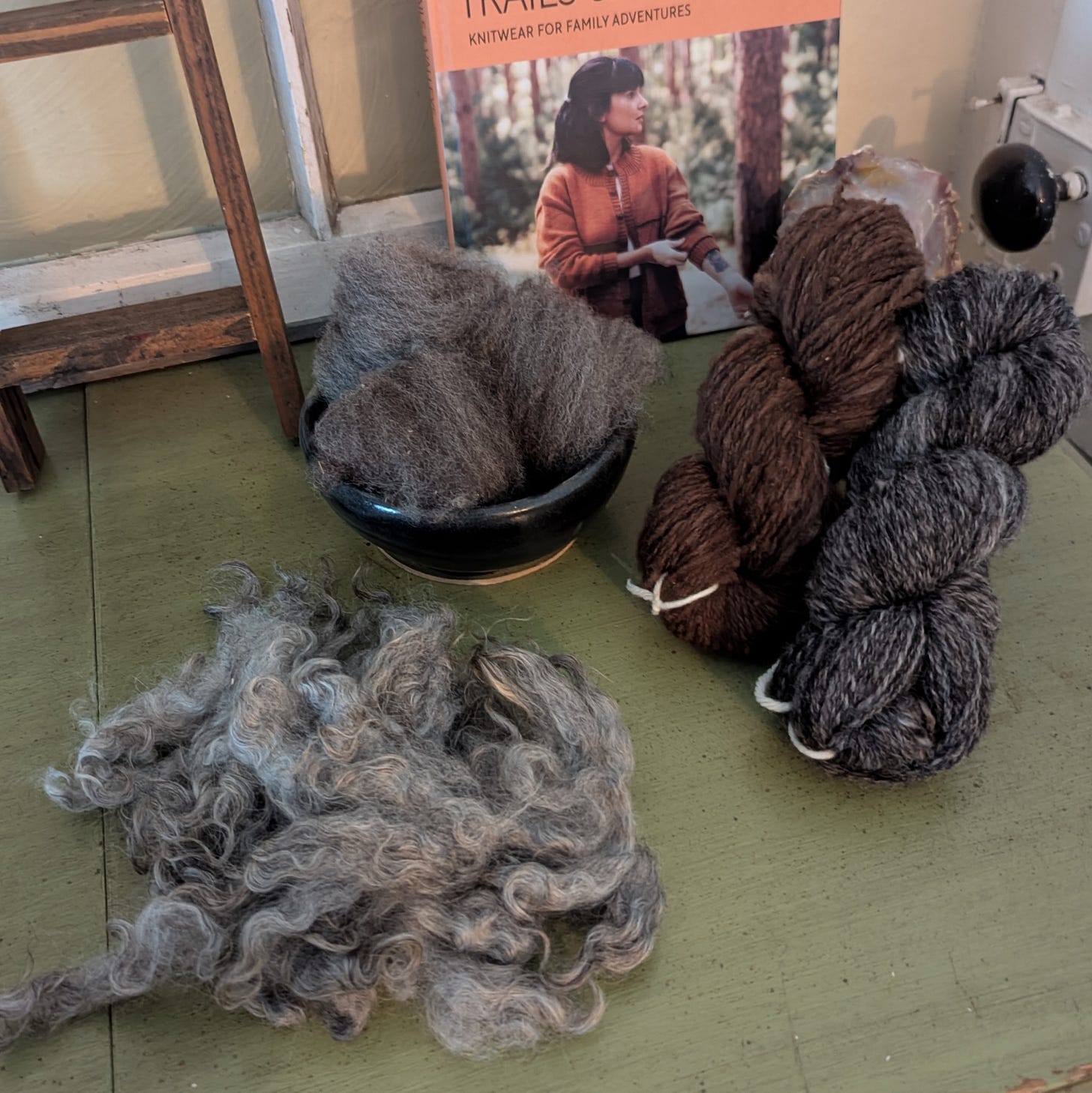 A small pile of washed Gotland wool locks sits on a table next to a small dark blue bowl filled with a few grey nests of combed Shetland wool. Two handspun skeins sit to the right of the prepped fiber: one chocolate brown Merino wool skein and one dark and light grey skein of Shetland wool yarn.