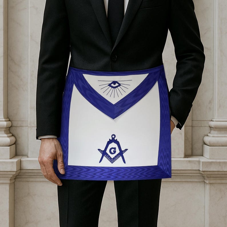 May include: A white apron with a blue border and accents. The apron features a design with an eye, sun rays, a compass, and a square. The apron is worn over a black suit. May include: A white apron with a blue border and accents. The apron features a design with an eye, sun rays, a compass, and a square. The apron is worn over a black suit.