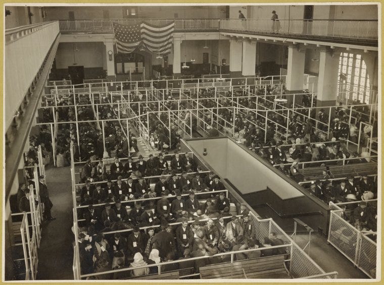 Black and white photo of the rectangular pens immigrants went through inside Ellis Island. A large space is divided into several spaces to the left and right of an open walkway in the center. Each pen contains lines of people.
