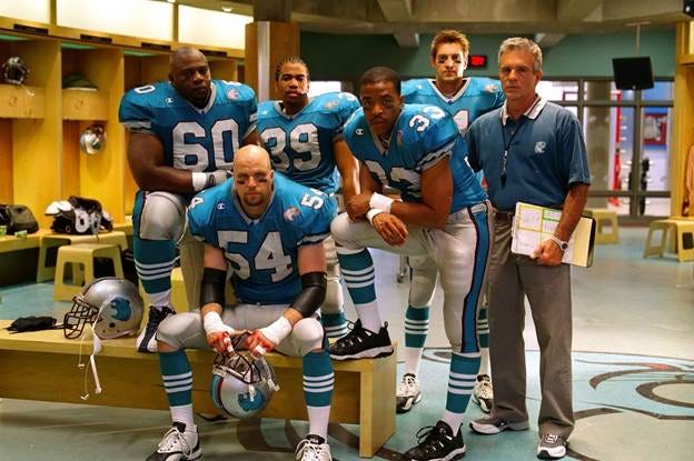 A photo of five Cougars football players in uniform in a locker room. The head coach stands next to them wearing a Cougars polo shirt and holding a clipboard. A photo of five Cougars football players in uniform in a locker room. The head coach stands next to them wearing a Cougars polo shirt and holding a clipboard.