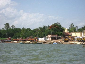 First image shows a traditional temple building with green and red striped walls a tall ornate pillar in front surrounded by trees and a clear blue sky. Second image depicts a long concrete bridge spanning over a wide river with calm dark water and distant green banks under a hazy sky. Third image captures a riverside scene with several boats docked along the muddy bank trees and small buildings including a temple with a dome in the background and construction equipment nearby. Fourth image features a red and yellow bridge extending over flowing brown water with railings people on motorcycles crossing and green vegetation on the sides.