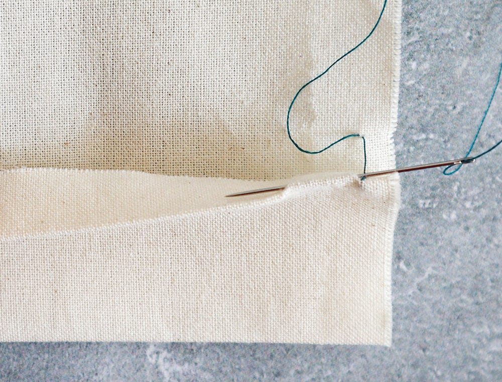 Cream muslin fabric with hem folded up, needle with teal thread sewing a slip stitch