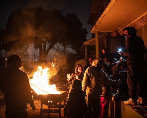 A group of people, most wearing hoodies, gathered around a fire in an outdoor barbecue grill outside a home at night. A group of people, most wearing hoodies, gathered around a fire in an outdoor barbecue grill outside a home at night.