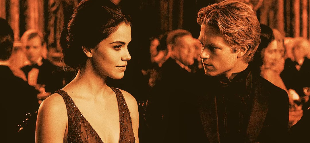 A dark-haired young woman in a glittering formal gown looks calmly toward a blond young man in black formal attire as they sit together at an elegant banquet surrounded by blurred guests and warm amber lighting.