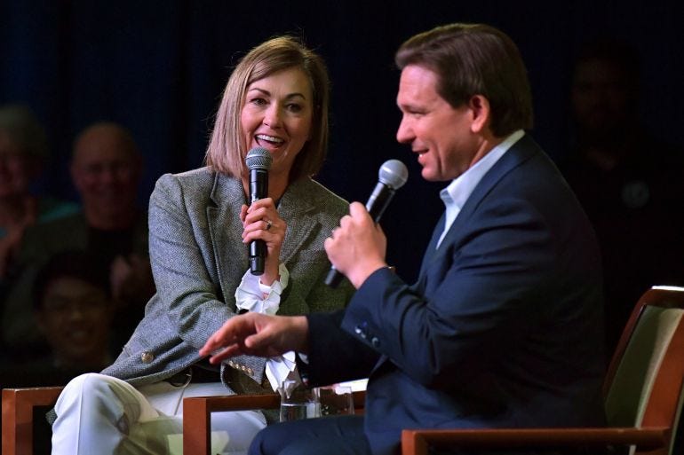 Kim Reynolds and Ron DeSantis sit side by side on a darkened stage, each holding a handheld microphone to their face as they speak. Kim Reynolds and Ron DeSantis sit side by side on a darkened stage, each holding a handheld microphone to their face as they speak.