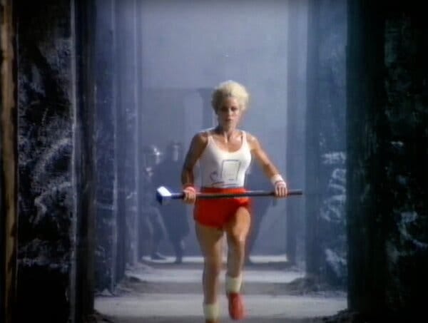 A woman with short white-blonde hair, a white tank top and red shorts runs through a corridor with a sledgehammer A woman with short white-blonde hair, a white tank top and red shorts runs through a corridor with a sledgehammer