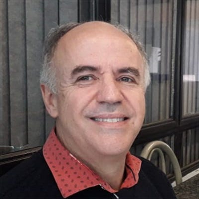 Homem calvo usando blusa de lã preta com o colarinho de uma camisa vermelha sobre a gola, sorrindo, sentado à esquerda de uma mesa de granito com uma xícara, um copo com café, um guardanapo e uma máscara, tendo um sofá de couro marrom do lado e um conjunto de janelas metálicas por trás Homem calvo usando blusa de lã preta com o colarinho de uma camisa vermelha sobre a gola, sorrindo, sentado à esquerda de uma mesa de granito com uma xícara, um copo com café, um guardanapo e uma máscara, tendo um sofá de couro marrom do lado e um conjunto de janelas metálicas por trás