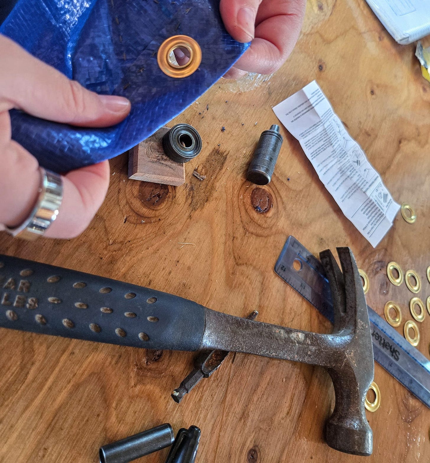 A close shot of hands holding an edge of blue tarp with a grommet installed. A hammer and grommet setting kit are in the background on the table.