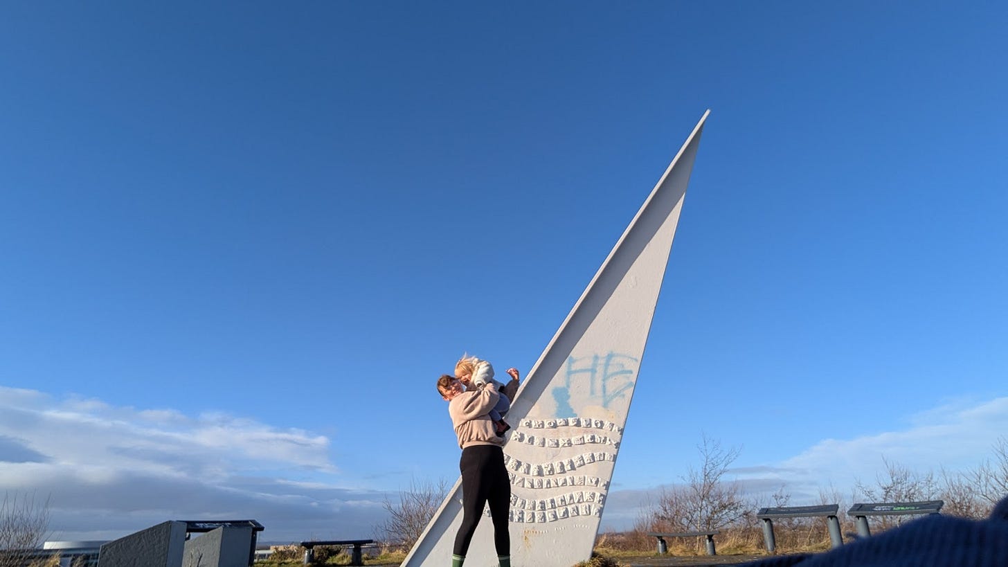 Self-timer photo of Ellen and her two-year old at the top of a big hill, standing in front of a sundial. The sky is blue and the sun is shining.
