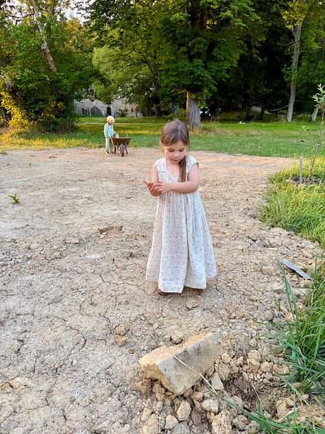Golden evening light on the château grounds: a child in a white nightgown walking toward the coach house past hay bales; a girl on a stone path with the château behind; a man and two children pushing a large round hay bale.