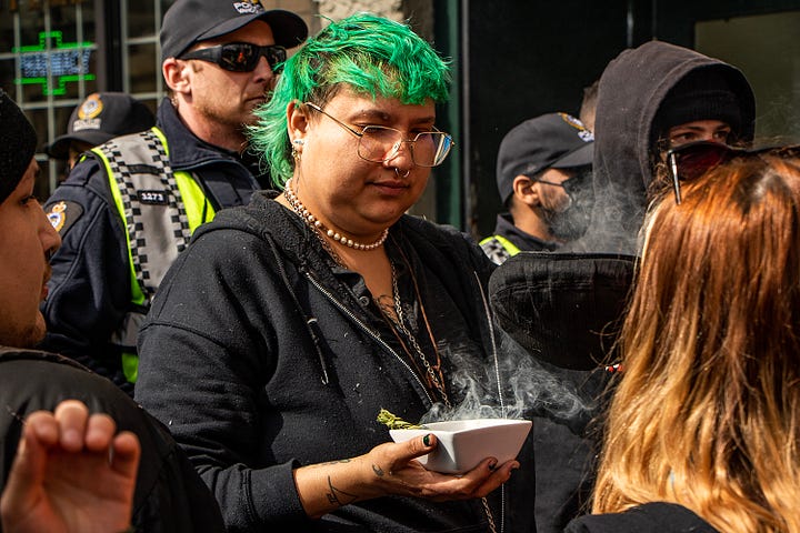 First image: A woman blows a cloud of vapour in the faces of police. A friend who was down there tells me her group was trolling the police the whole time, including taking drags from their vape and blowing it at the police. Second image: A person with a white shirt and a white wrap around their head and face and a black cap holds up a sign that reads: "Cops are better at committing crimes than solving them." Third image: An Indigenous person, back facing the line of police, is holding a bowl of smoking sage. After holding a cap overtop of the sage smoke, they turned the cap over, and smoke can be seen trickling upwards, out of the hat. Fourth image: A couple of police officers are seen at the front line. One is wearing a mask and looking dead ahead. The other is looking at something to the left of the camera's perspective.