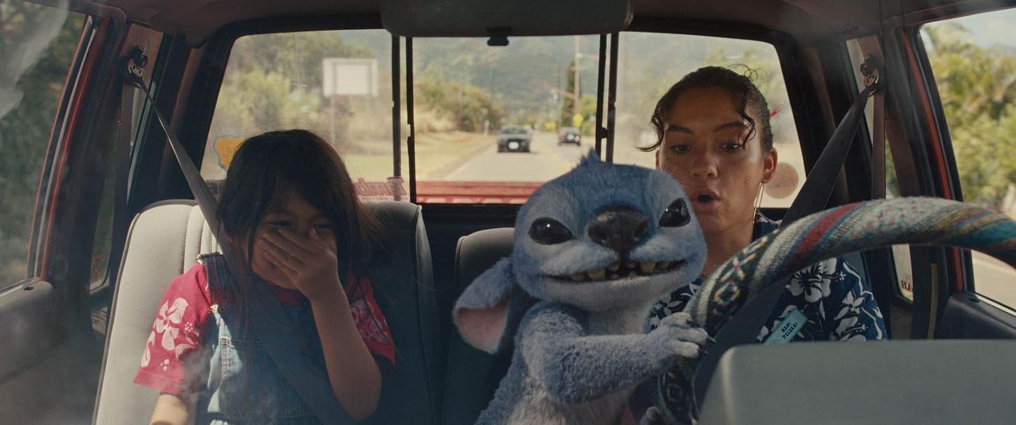 A little girl (Lilo) and her older sister (Nani) are in a pickup truck where a blue fluffy alien (Stitch) is taking the wheel away from Nani. Lilo laughs while Nani is shocked.