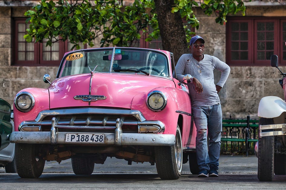 A Havana cab driver in sunglasses leans against his vintage pink convertible taxi.