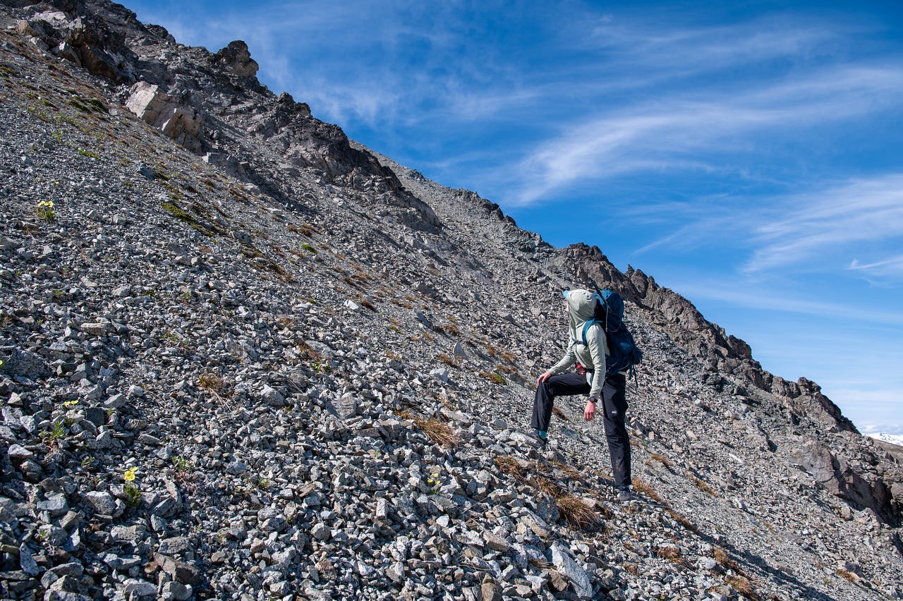 A hiker in a pale hooded shirt and dark pants leans into a steep slope of loose grey scree, a hand on one knee, climbing beside a dark rock rib. A few small yellow flowers grow among the rocks, and thin cloud streaks the deep blue sky above.
