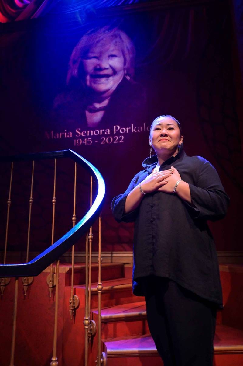 a Filipina woman in black stands on a stage with her hands over her heart in front of a large projected image of her grandmother a Filipina woman in black stands on a stage with her hands over her heart in front of a large projected image of her grandmother