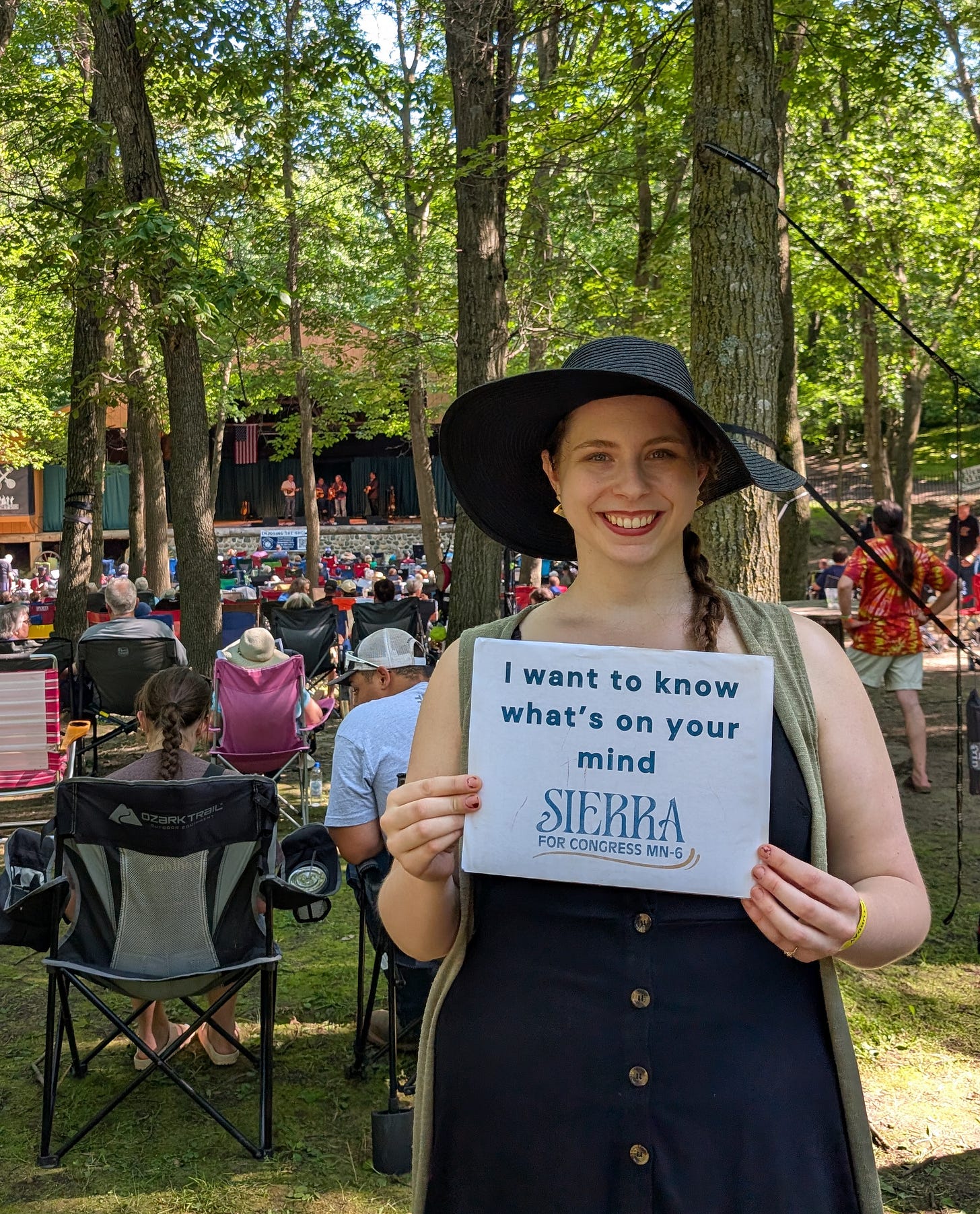Sierra Grandy in front of a crowd at a music venue. Sierra is a white women in a black dress and black hat, holding a sign that says "I want to know what's on your mind." with a logo that says "Sierra for Congress MN-6"