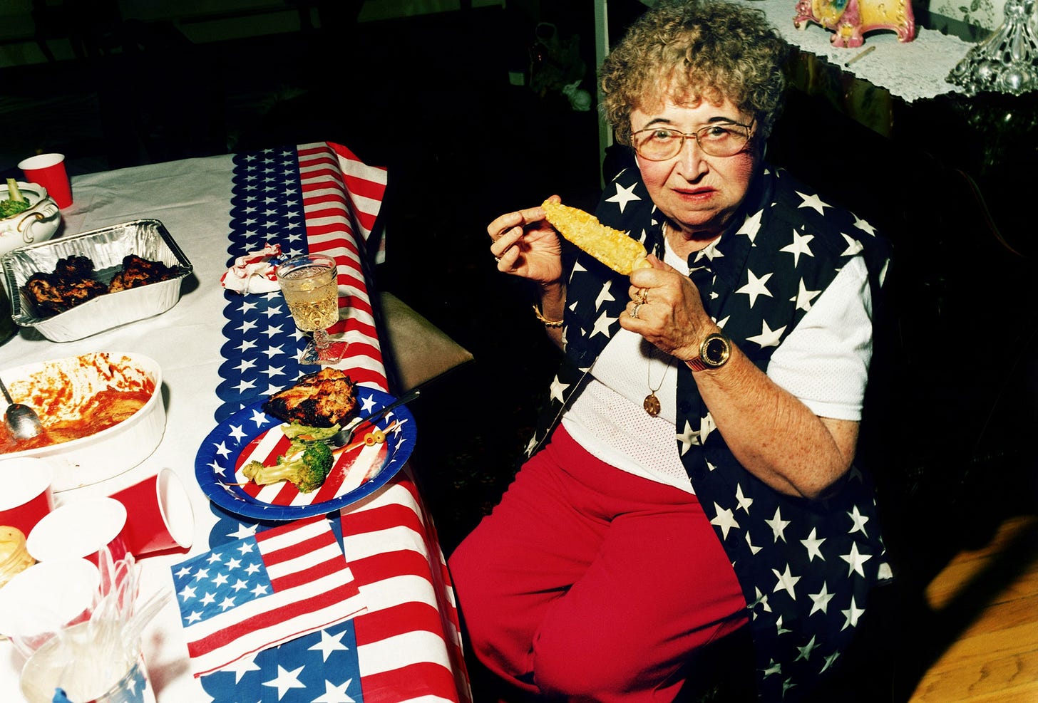 A photo of an older woman eating a corn on the cob while wearing American flagthemed clothes.
