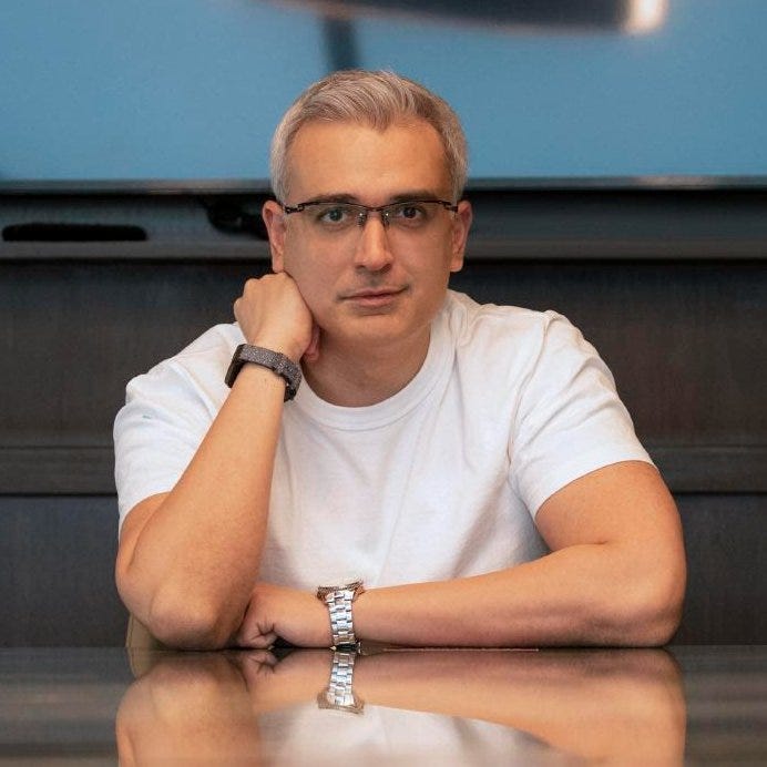 A photo of Pav Gill at a mirrored table with this head resting on his hand, one arm on the table. He has short grey hair and is wearing a white t-shirt and glasses.
